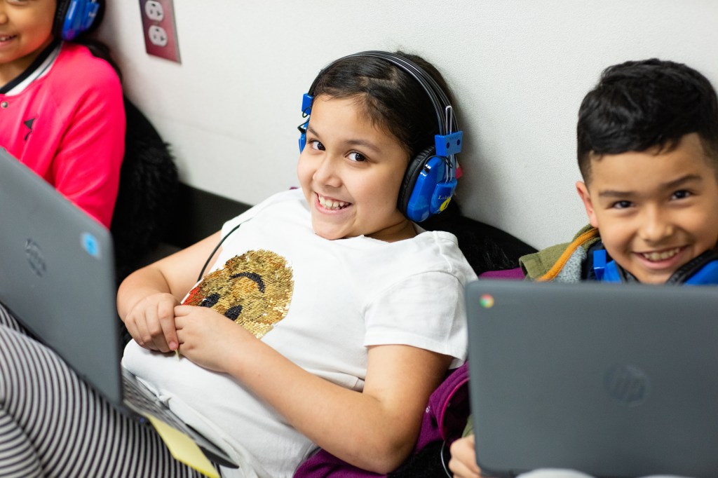 A student wearing headphones with a laptop in front of her smiles at the camera. A student next to her with a laptop also smiles for the camera.